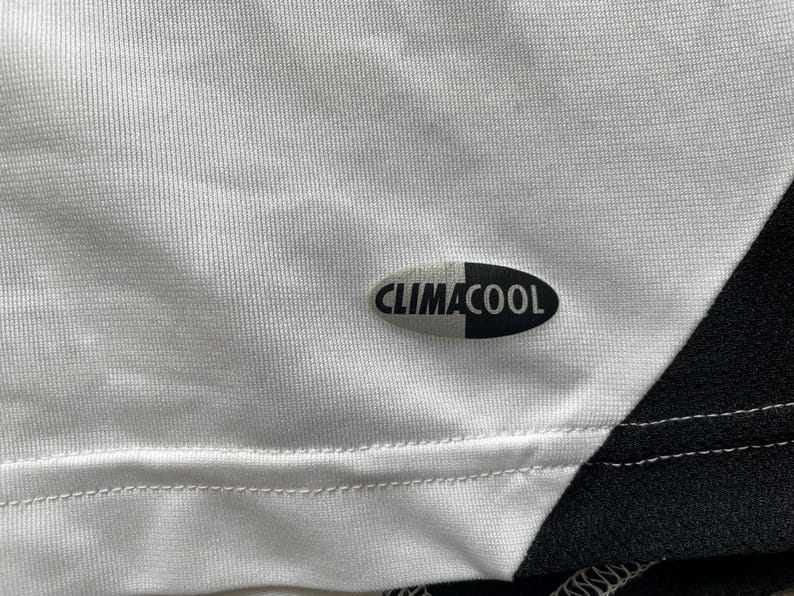 Pode incluir: Close-up de uma camisa atl&eacute;tica branca com um design em preto e branco. A camisa apresenta o logotipo "CLIMACOOL" em preto e branco. O tecido tem uma apar&ecirc;ncia texturizada.