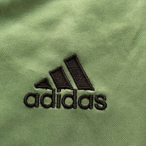 May include: Close-up of a green athletic fabric with the embroidered Adidas logo in black. The fabric has a textured appearance, with a black and white stripe at the top.