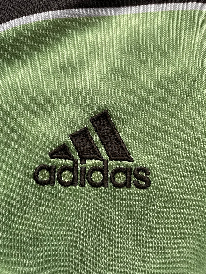 May include: Close-up of a green athletic fabric with the embroidered Adidas logo in black. The fabric has a textured appearance, with a black and white stripe at the top.