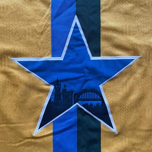 May include: Close-up of a gold-colored fabric with a repeating pattern. A blue star with a white outline is centered, containing a black silhouette of a cityscape. Vertical stripes of blue and green are behind the star.