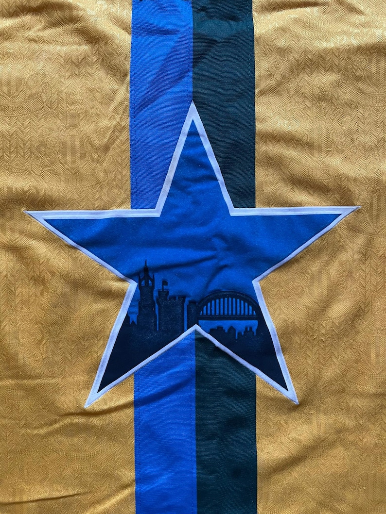May include: Close-up of a gold-colored fabric with a repeating pattern. A blue star with a white outline is centered, containing a black silhouette of a cityscape. Vertical stripes of blue and green are behind the star.