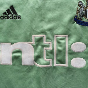 May include: Close-up of a light green soccer jersey with the Adidas logo in black. The jersey features the Newcastle United crest and the embroidered text "nti:" in white.