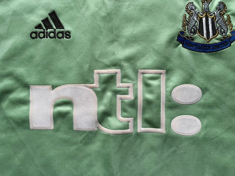 May include: Close-up of a light green soccer jersey with the Adidas logo in black. The jersey features the Newcastle United crest and the embroidered text "nti:" in white.