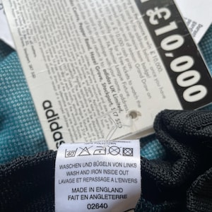 K&ouml;nnte beinhalten: Nahaufnahme eines Adidas-Etiketts mit Waschanleitung auf Deutsch, Englisch und Franz&ouml;sisch sowie dem Text "Made in England". Ein zweites Etikett wirbt f&uuml;r die Chance auf 10.000 &pound;.