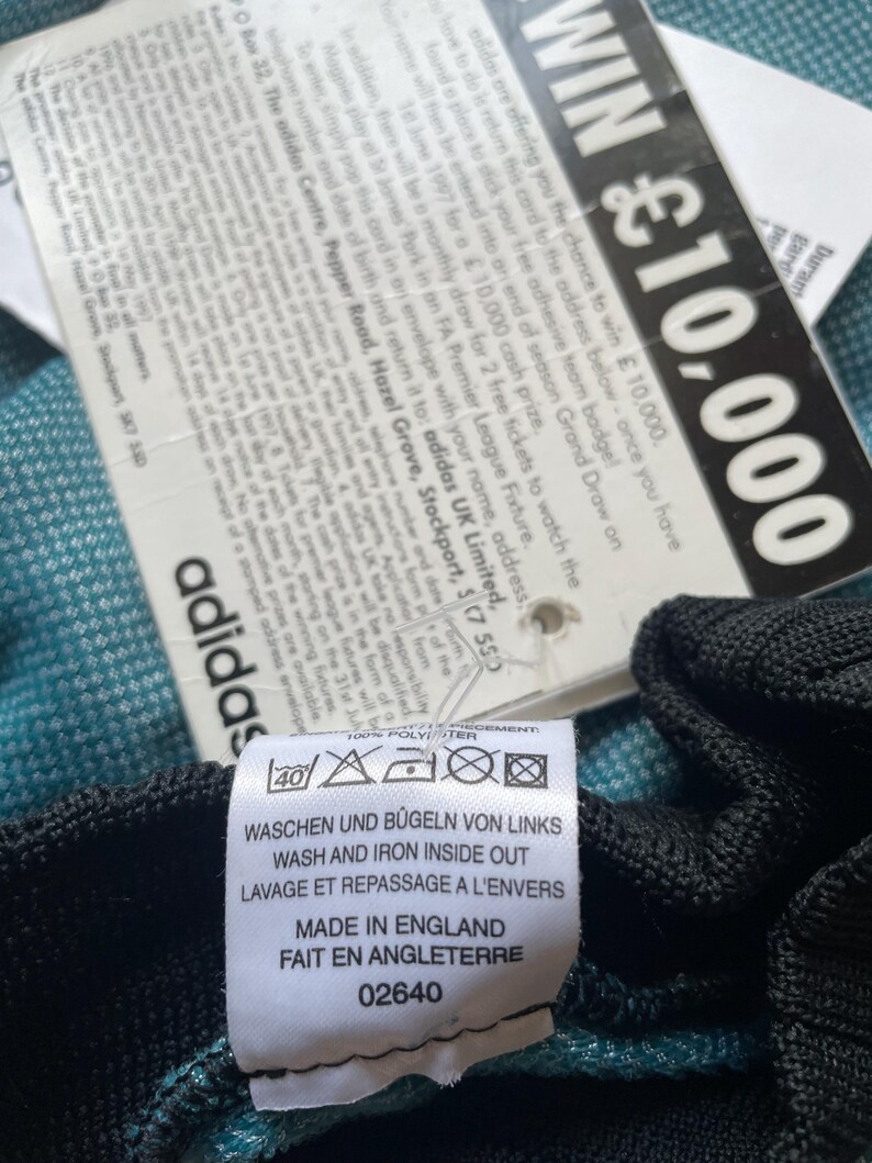 K&ouml;nnte beinhalten: Nahaufnahme eines Adidas-Etiketts mit Waschanleitung auf Deutsch, Englisch und Franz&ouml;sisch sowie dem Text "Made in England". Ein zweites Etikett wirbt f&uuml;r die Chance auf 10.000 &pound;.