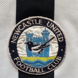 Może przedstawiać: Zbliżenie naszywki Newcastle United Football Club. Naszywka przedstawia srokę, dwa niebieskie zamki i napisy "Newcastle United Football Club" w okrągłym wzorze. Tło to czarno-biała tkanina w paski.
