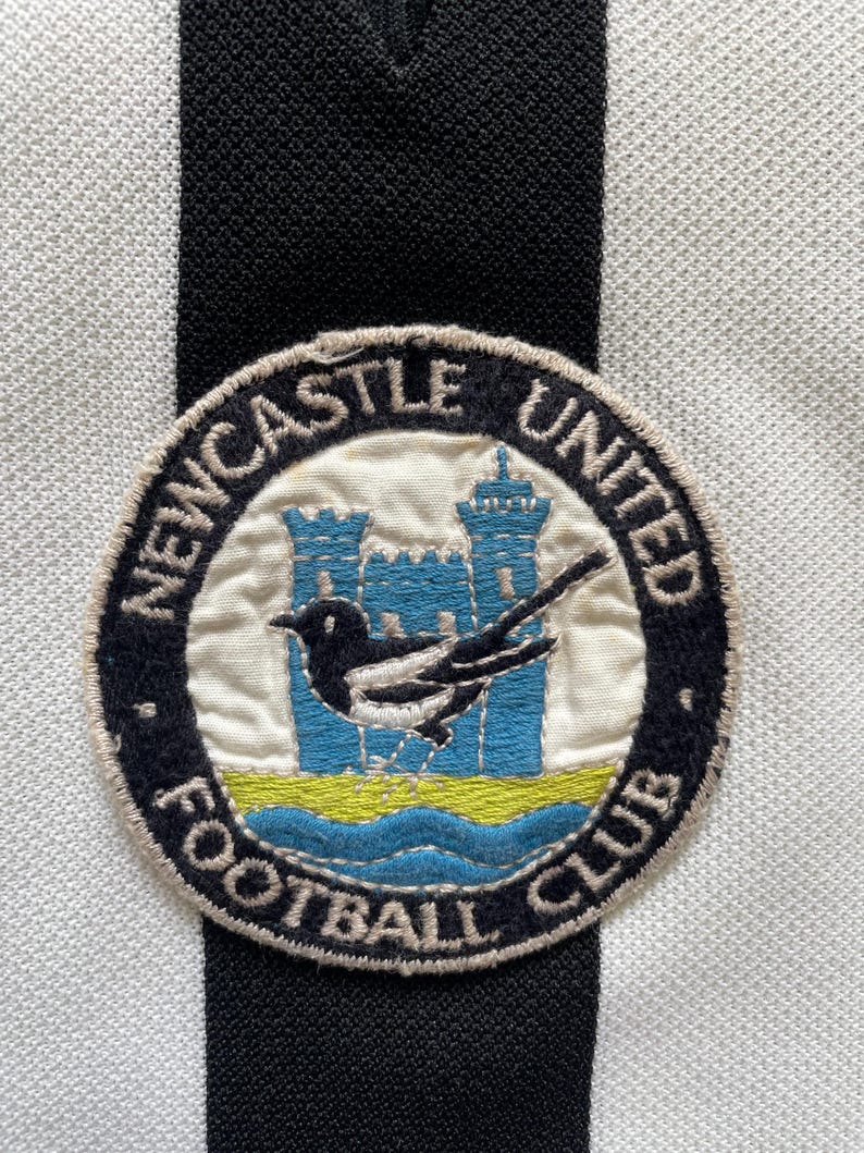 Może przedstawiać: Zbliżenie naszywki Newcastle United Football Club. Naszywka przedstawia srokę, dwa niebieskie zamki i napisy "Newcastle United Football Club" w okrągłym wzorze. Tło to czarno-biała tkanina w paski.