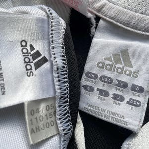 May include: White and black Adidas soccer jersey with black stripes. The jersey has a white tag with the Adidas logo and the text "The brand with the 3 stripes" and "Die Weltmarke mit den 3 stripes". The tag also has the text "04/05 110156 AH JOO".