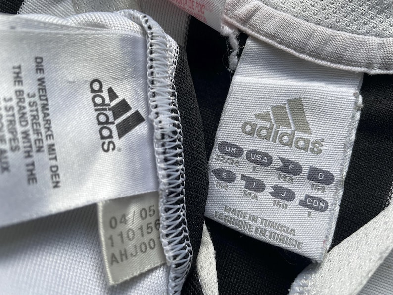 May include: White and black Adidas soccer jersey with black stripes. The jersey has a white tag with the Adidas logo and the text "The brand with the 3 stripes" and "Die Weltmarke mit den 3 stripes". The tag also has the text "04/05 110156 AH JOO".