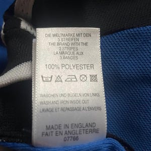 K&ouml;nnte beinhalten: Nahaufnahme eines Etiketts mit Text in Englisch, Deutsch und Franz&ouml;sisch. Das Etikett lautet "100% POLYESTER" und enth&auml;lt Pflegehinweise. Das Etikett ist an einem blauen und schwarzen Stoff befestigt.