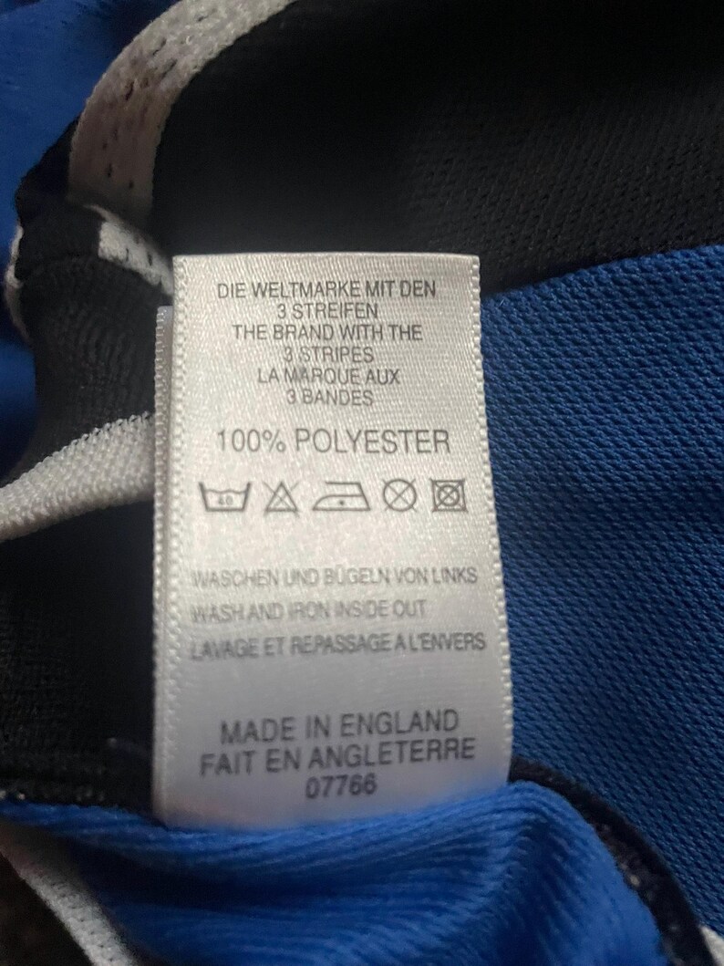 K&ouml;nnte beinhalten: Nahaufnahme eines Etiketts mit Text in Englisch, Deutsch und Franz&ouml;sisch. Das Etikett lautet "100% POLYESTER" und enth&auml;lt Pflegehinweise. Das Etikett ist an einem blauen und schwarzen Stoff befestigt.