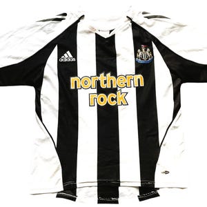 May include: A white and black striped soccer jersey with the Adidas logo on the left chest and the Newcastle United crest on the right chest. The jersey has "northern rock" written in gold letters across the chest.