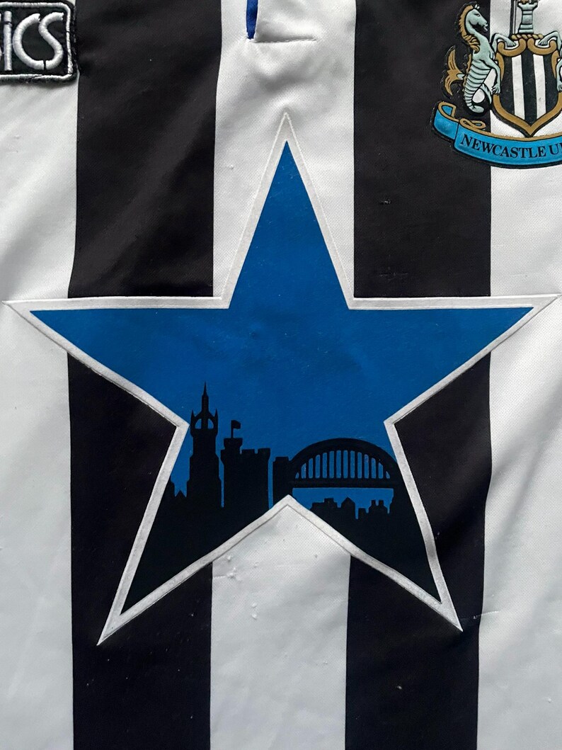 May include: A black and white striped jersey with a blue star-shaped emblem. The emblem features a silhouette of a city skyline. The jersey also has a team logo and the word "ICS" on the upper left.