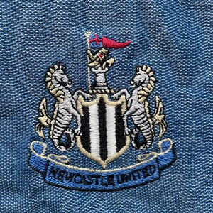 May include: Embroidered Newcastle United crest patch. The design features a shield with black and white stripes, flanked by seahorses, and a blue banner with "Newcastle United" on a denim blue background.