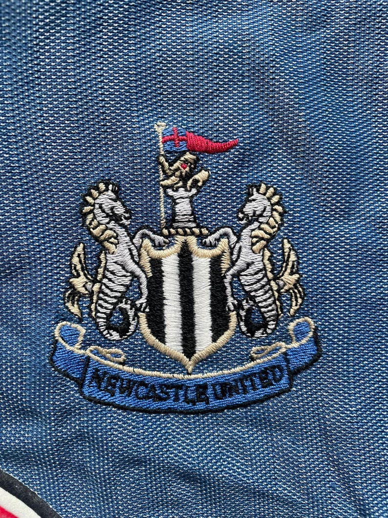 May include: Embroidered Newcastle United crest patch. The design features a shield with black and white stripes, flanked by seahorses, and a blue banner with "Newcastle United" on a denim blue background.