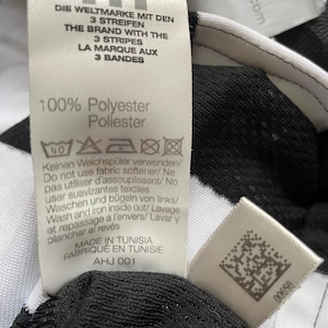 May include: A close-up of a black and white Adidas sports garment tag. The tag includes text in multiple languages: English, German, and French. The tag also states the garment is 100% polyester and made in Tunisia.