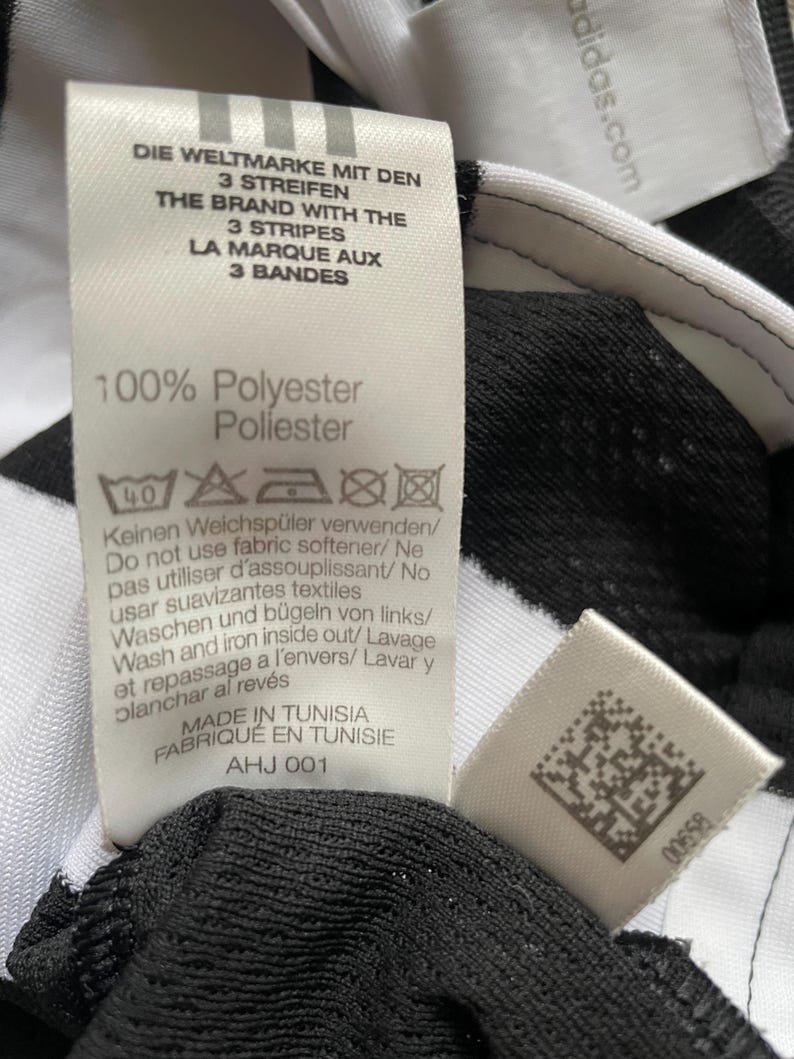 May include: A close-up of a black and white Adidas sports garment tag. The tag includes text in multiple languages: English, German, and French. The tag also states the garment is 100% polyester and made in Tunisia.
