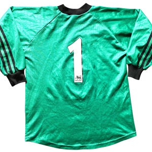 K&ouml;nnte beinhalten: Lang&auml;rmeliges, smaragdgr&uuml;nes Fu&szlig;balltrikot mit schwarzen Akzenten. Auf der R&uuml;ckseite ist die wei&szlig;e Nummer 1 zu sehen. Das Trikot hat schwarze Streifen auf den &Auml;rmeln, einen schwarzen Kragen und B&uuml;ndchen und ein strukturiertes Muster.