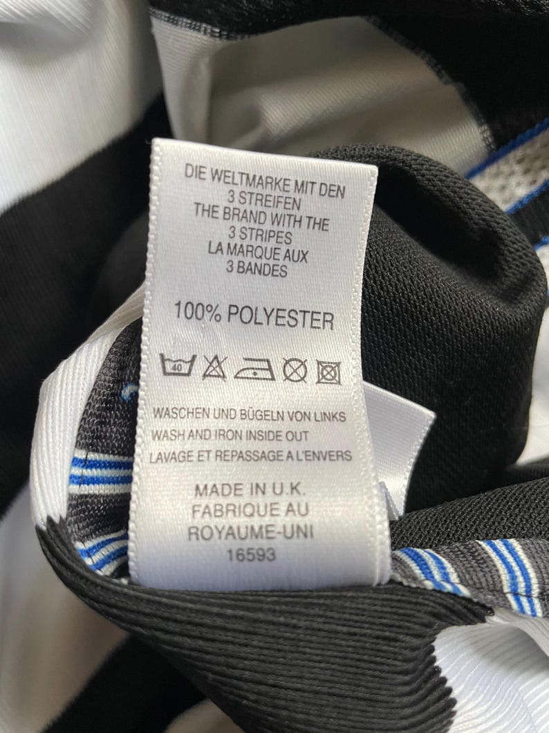 May include: White and black striped shirt tag with care instructions and information about the brand. The tag says "Die Weltmarke mit den 3 Streifen", "The Brand with the 3 Stripes", "La Marque aux 3 Bandes", "100% Polyester", "Waschen und b&uuml;geln von links", "Wash and iron inside out", "Lavage et repassage a l'envers", "Made in U.K.", "Fabrique au Royaume-Uni", and "16593".
