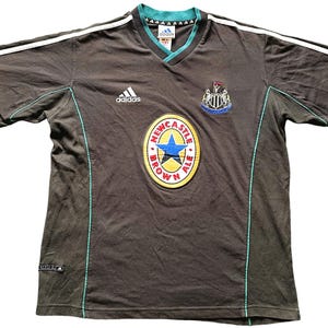 May include: A black short-sleeved t-shirt with white stripes on the sleeves and a green trim. The shirt has an Adidas logo on the left chest and a Newcastle Brown Ale logo on the front. The Newcastle Brown Ale logo is a round, yellow circle with a blue star and the words "Newcastle Brown Ale" in red.