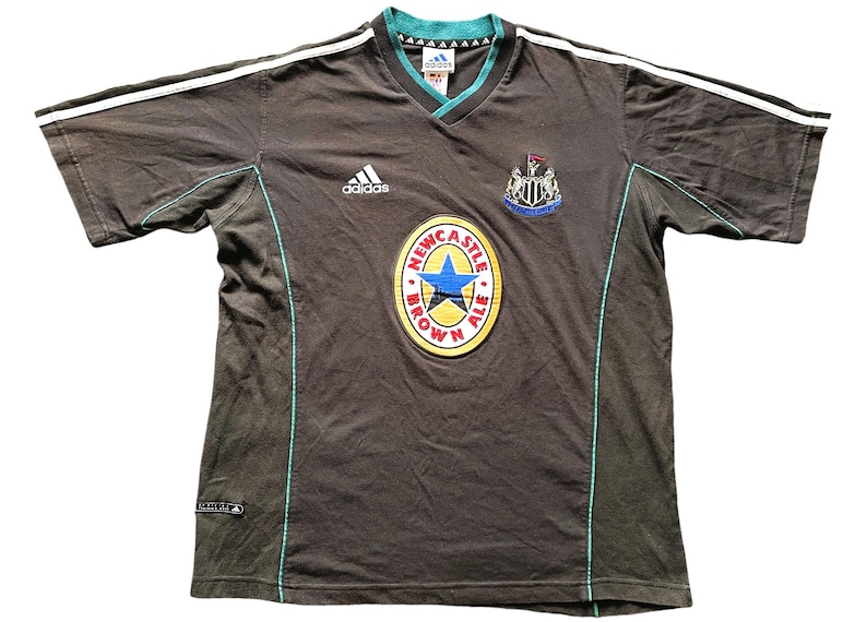 May include: A black short-sleeved t-shirt with white stripes on the sleeves and a green trim. The shirt has an Adidas logo on the left chest and a Newcastle Brown Ale logo on the front. The Newcastle Brown Ale logo is a round, yellow circle with a blue star and the words "Newcastle Brown Ale" in red.