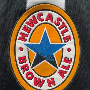 May include: Close-up of a Newcastle Brown Ale patch. The patch is oval-shaped with a blue star in the center, surrounded by the words "Newcastle Brown Ale" in red. The background is black and white striped.