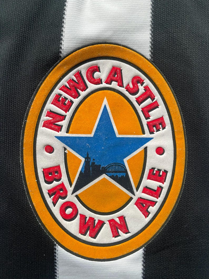 May include: Close-up of a Newcastle Brown Ale patch. The patch is oval-shaped with a blue star in the center, surrounded by the words "Newcastle Brown Ale" in red. The background is black and white striped.