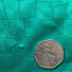 May include: A silver 50 pence coin featuring a seated Britannia figure, set against a textured green fabric background. The coin is a heptagonal shape, with the text "FIFTY PENCE" and "50" visible.