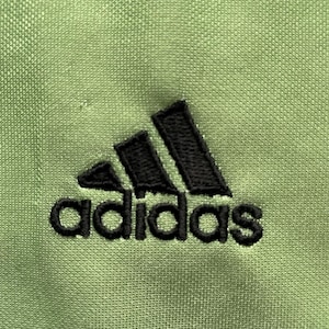 Pu&ograve; includere: Primo piano di un tessuto verde chiaro con il logo Adidas nero ricamato sul davanti. Il logo presenta tre strisce nere sopra la parola "adidas" in nero, con un carattere senza grazie.