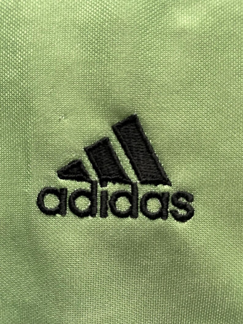 Pu&ograve; includere: Primo piano di un tessuto verde chiaro con il logo Adidas nero ricamato sul davanti. Il logo presenta tre strisce nere sopra la parola "adidas" in nero, con un carattere senza grazie.