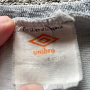 May include: White tag with the text "The Choice of Champions" and the logo for the brand "Umbro" in orange. The tag is attached to a gray shirt.