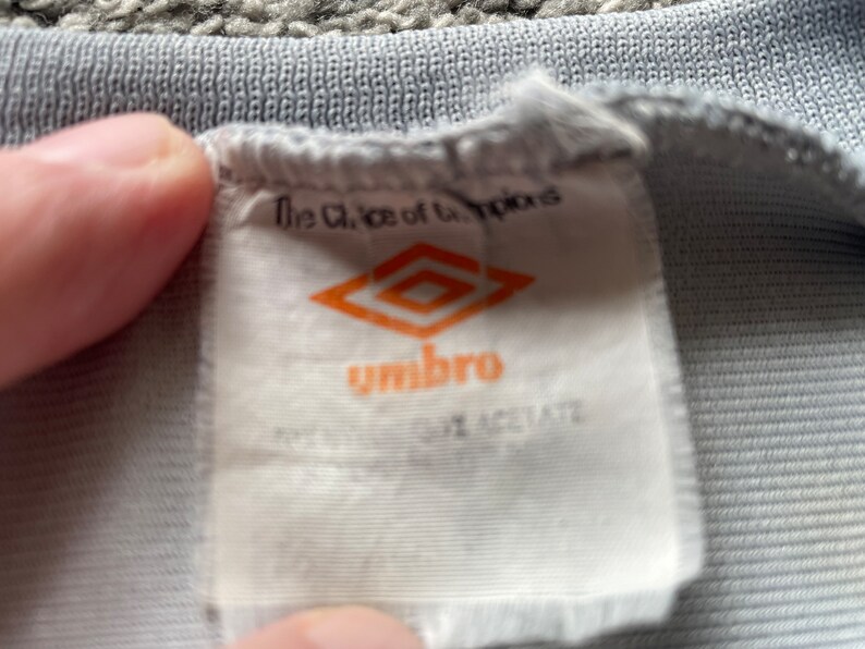 May include: White tag with the text "The Choice of Champions" and the logo for the brand "Umbro" in orange. The tag is attached to a gray shirt.