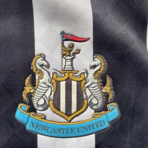 Op de afbeelding: Close-up van een zwart-wit gestreept voetbalshirt met het Newcastle United-embleem. Het embleem bevat twee zeepaarden, een schild met verticale strepen en een banner met de tekst "Newcastle United".