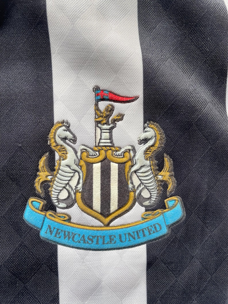 Op de afbeelding: Close-up van een zwart-wit gestreept voetbalshirt met het Newcastle United-embleem. Het embleem bevat twee zeepaarden, een schild met verticale strepen en een banner met de tekst "Newcastle United".