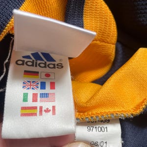 May include: A white tag with the Adidas logo and text, along with a row of international flags. The tag is attached to a navy blue and yellow striped shirt. The tag also includes the numbers 971001 and 06/01.