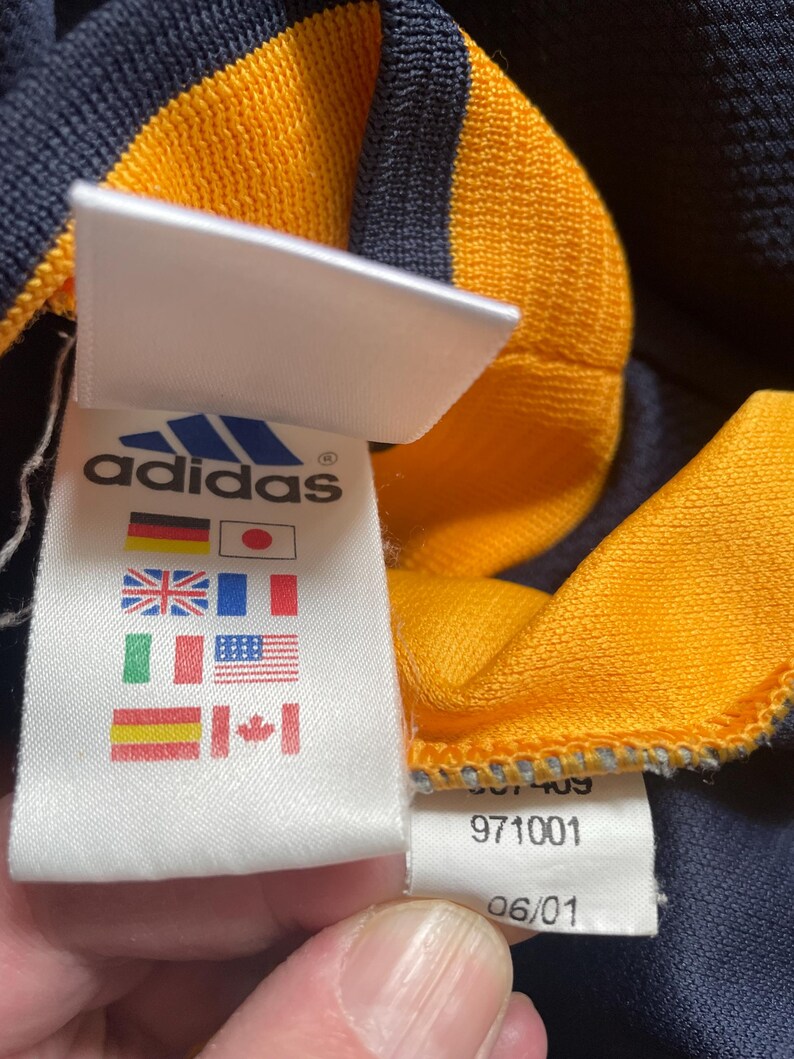 May include: A white tag with the Adidas logo and text, along with a row of international flags. The tag is attached to a navy blue and yellow striped shirt. The tag also includes the numbers 971001 and 06/01.