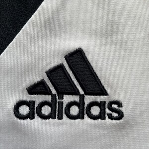 Pode incluir: Close-up de um tecido branco com o logotipo Adidas preto bordado. O logotipo Adidas &eacute; um design de tri&acirc;ngulo preto com a palavra "adidas" bordada abaixo em preto.