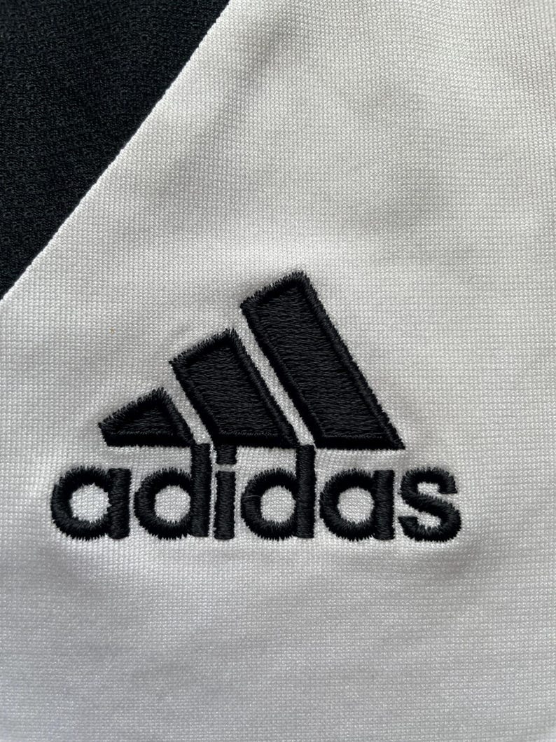 Pode incluir: Close-up de um tecido branco com o logotipo Adidas preto bordado. O logotipo Adidas &eacute; um design de tri&acirc;ngulo preto com a palavra "adidas" bordada abaixo em preto.