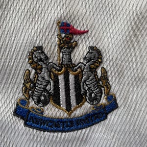 Pu&ograve; includere: Una toppa ricamata bianca con uno scudo a strisce bianche e nere, due ippocampi dorati e una corona dorata. Lo scudo ha una bandiera bianca e nera in cima. Il testo "NEWCASTLE UNITED" &egrave; su un nastro blu.