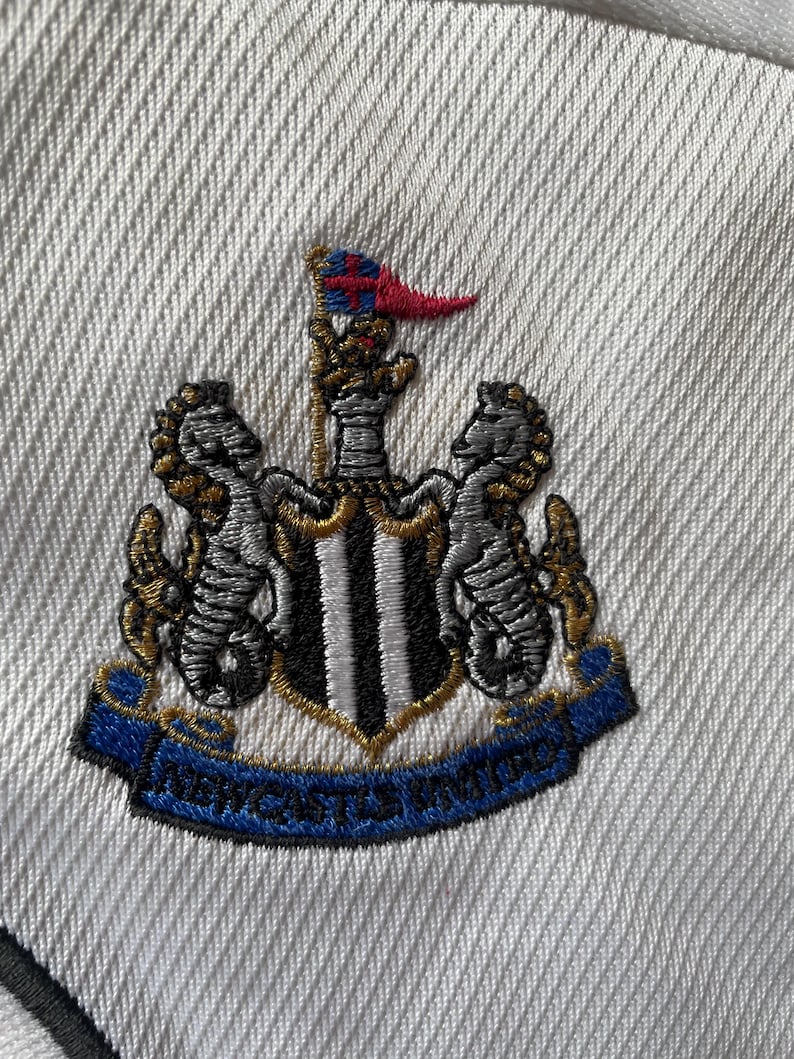 Pu&ograve; includere: Una toppa ricamata bianca con uno scudo a strisce bianche e nere, due ippocampi dorati e una corona dorata. Lo scudo ha una bandiera bianca e nera in cima. Il testo "NEWCASTLE UNITED" &egrave; su un nastro blu.