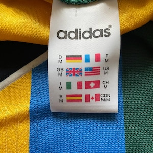 May include: Close-up of an Adidas tag on a jersey. The tag displays the Adidas logo and country flags, including the US flag. The jersey has yellow, blue, and green sections.
