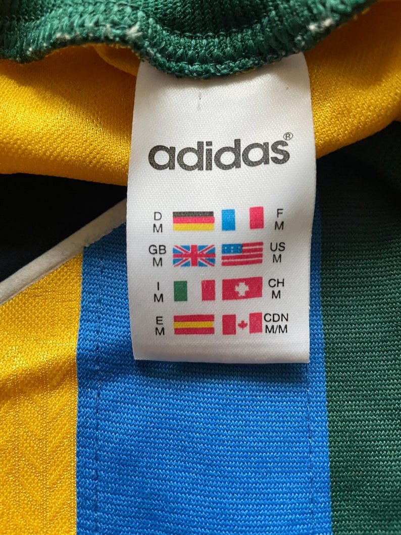 May include: Close-up of an Adidas tag on a jersey. The tag displays the Adidas logo and country flags, including the US flag. The jersey has yellow, blue, and green sections.