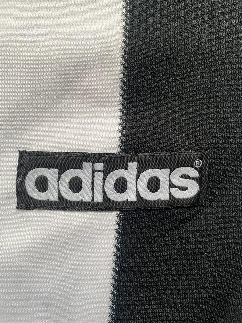 May include: Close-up of a black and white athletic jersey. The jersey is divided vertically, with a white section on the left and a black section on the right. The Adidas logo is prominently displayed on a black patch.