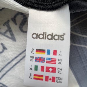 May include: A close-up of an Adidas clothing label. The white label shows the Adidas logo and size details with flags representing different countries, including the UK, and Canadian flags. The label is attached to a white and black garment.