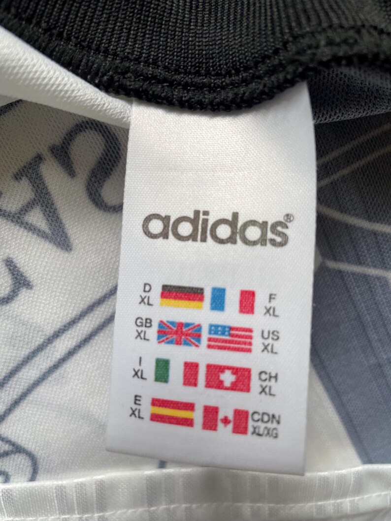 May include: A close-up of an Adidas clothing label. The white label shows the Adidas logo and size details with flags representing different countries, including the UK, and Canadian flags. The label is attached to a white and black garment.