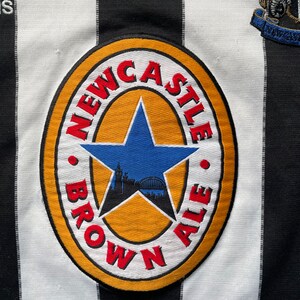 Może przedstawiać: Czarno-biała, pasiasta koszulka piłkarska z logo Newcastle Brown Ale z przodu. Logo to ż&oacute;łte koło z czerwoną gwiazdą i napisem "Newcastle Brown Ale" wok&oacute;ł krawędzi.