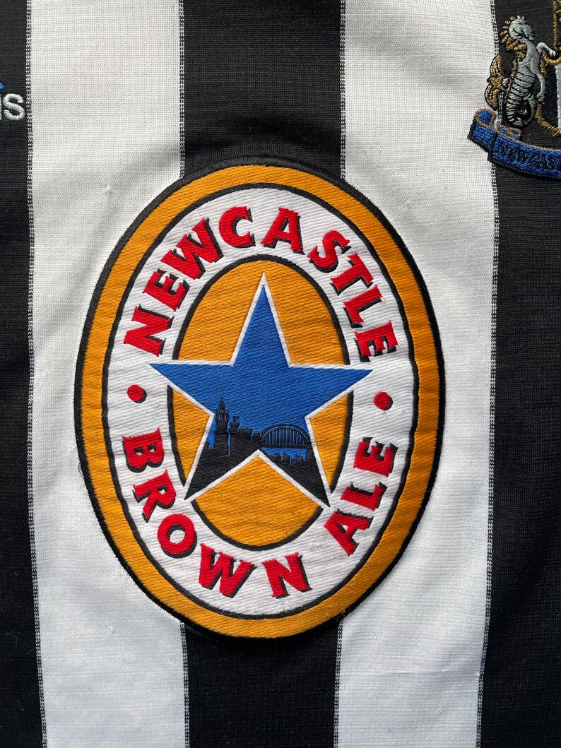 Może przedstawiać: Czarno-biała, pasiasta koszulka piłkarska z logo Newcastle Brown Ale z przodu. Logo to ż&oacute;łte koło z czerwoną gwiazdą i napisem "Newcastle Brown Ale" wok&oacute;ł krawędzi.