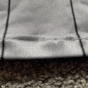 May include: Close-up of a gray fabric with black stripes. The fabric has a sewn edge and a textured surface.