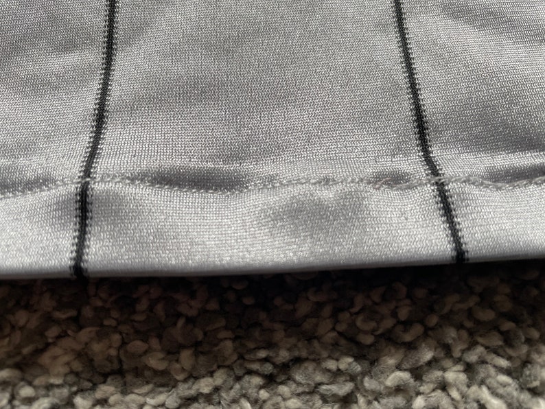 May include: Close-up of a gray fabric with black stripes. The fabric has a sewn edge and a textured surface.