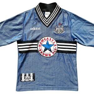 May include: A vintage blue soccer jersey with black and white accents. The jersey features the Adidas logo, a Newcastle United crest, and a "Newcastle Brown Ale" patch. The collar and sleeves have black and white stripes.